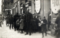 Schwarz-Weiß-Photographie: Schlange von Menschen vor Geschäft, von zwei Uniformierten begleitet