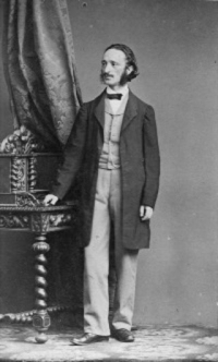 Julius Rodenberg Schwarz-Weiß-Portraitphotographie: Mann mit halblangen Haaren & Schnauzbart steht, Hand auf Tisch