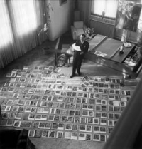 André Malraux inmitten der Abbildungen für Das imaginäre Museum um 1950. Aus: Rosalind Krauss: "Das Schicksalsministerium". In: Paradigma Fotografie. Fotokritik am Ende des fotografischen Zeitalters (2002), S.395 Schwarz-Weiß-Foto: stehender Mann mit Stück Papier in der Hand, auf dem Boden zahlreiche Bilder